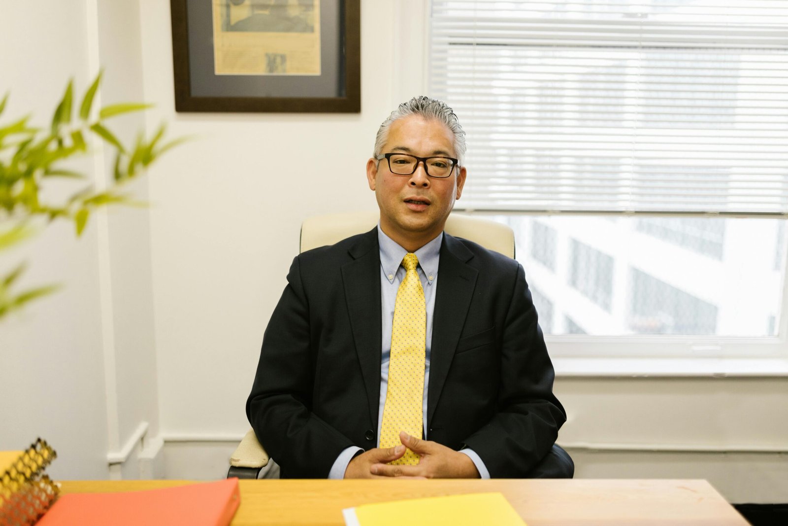 Middle-aged man in business attire sitting at an office desk with a blurred background.